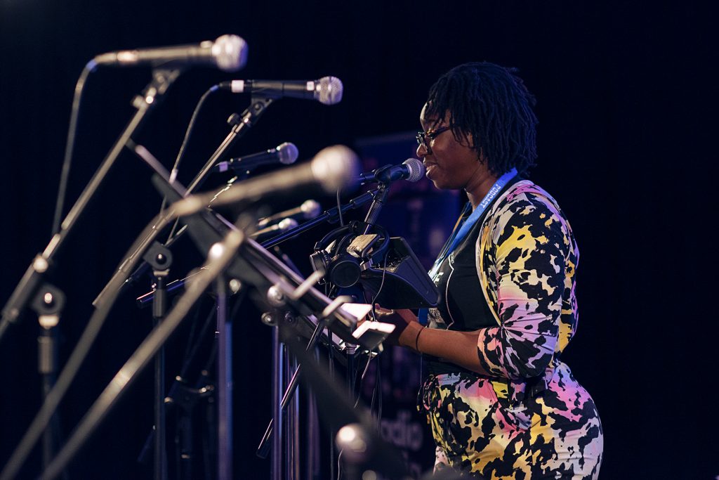 Aminita Francis performing as Lenses at the London Podcast Festival for Radio Elusia Live. Aminita is wearing a pink, yellow, black and white camouflage like outfit and is stood behind a row of microphones.