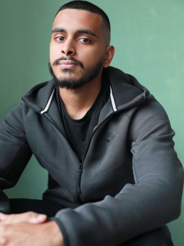 A headshot of a south Asian actor wearing a dark grey hoodie, with dark brown shaved hair